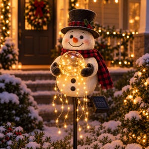 Könnte beinhalten: Ein Schneemann-Gartenstecker mit texturiertem weißem Körper, schwarzem Zylinder mit rot-kariertem Band und Stechpalme sowie einem rot-schwarz karierten Schal. Der Schneemann hält eine klare Kugel mit warmweißen Lichterketten und einem Solarpanel.