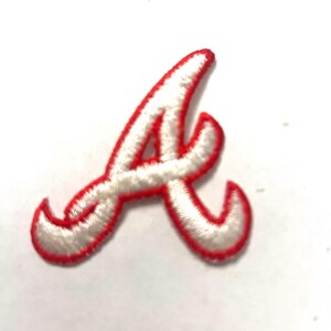 May include: An embroidered patch in the shape of a stylized capital letter A. The letter is white with a thick, vibrant red border, featuring a cursive-inspired design with pointed ends and a distinct, sweeping stroke characteristic of sports logos.