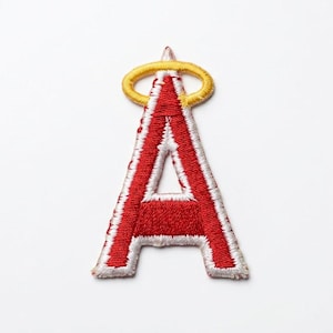 May include: An embroidered patch in the shape of the capital letter A, featuring a vibrant red center with a thick white border. A yellow halo is stitched directly above the top point of the letter, completing the angel-themed design.