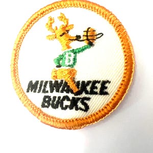 Puede incluir: Un parche bordado circular con fondo blanco y borde naranja. Presenta una mascota de ciervo amarilla con una camiseta verde con una letra B blanca, sosteniendo un balón de baloncesto. El texto MILWAUKEE BUCKS está bordado en color negro.