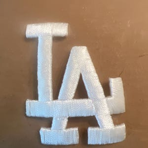 May include: A white embroidered patch featuring the interlocking LA logo. The design consists of the capital letters L and A connected in a stylized script, with a dense, textured thread finish throughout the entire surface of the emblem.