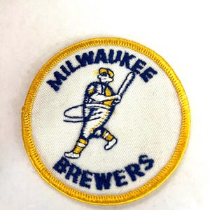 May include: A circular embroidered patch with a white background and a yellow border. It features a blue and yellow baseball player graphic in the center, with the blue text MILWAUKEE arched above and BREWERS arched below the figure.