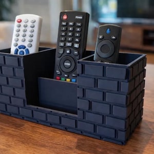 May include: A dark gray rectangular remote control holder with a textured brick pattern on the exterior. The organizer features three distinct compartments of varying heights, currently holding one white and two black remote controls.