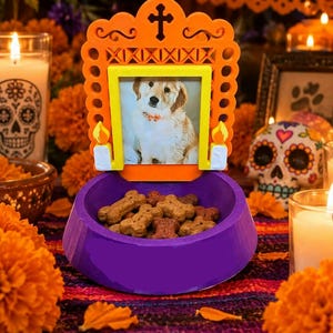 May include: A purple pet bowl with an attached orange frame featuring a cross and two candle motifs. The frame holds a photograph of a light-colored puppy wearing a bow tie. The bowl is filled with brown bone-shaped dry pet food treats.