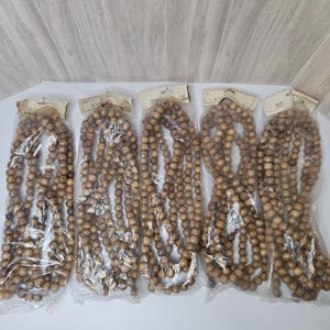 May include: Five individual strands of round, light brown wooden beads, each enclosed in a clear plastic bag with a paper header card attached at the top. The beads are uniform in size and color, arranged in long, continuous loops within the packaging.