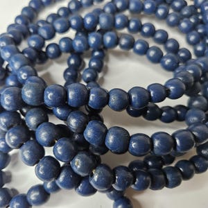 May include: A collection of round, dark blue beads strung together on a thin cord. The beads have a smooth, matte finish and are uniform in size, creating a continuous, tangled loop of deep navy blue spheres that are arranged in a dense cluster.
