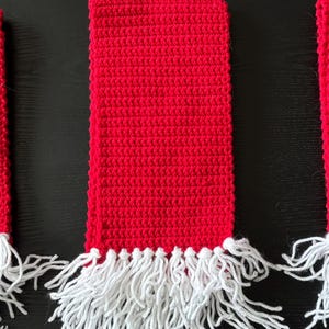 May include: Three rectangular scarves crocheted in a vibrant red yarn. Each scarf features a consistent textured stitch pattern throughout the length and is finished with a thick fringe of white yarn tassels attached to the bottom edge.