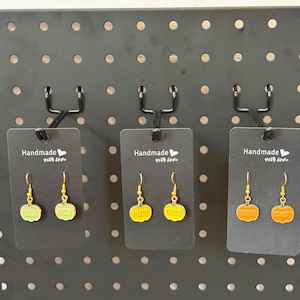 May include: Three pairs of dangle earrings on black display cards. The first pair features light green pumpkins with the word GRATEFUL, the second pair features yellow pumpkins with BLESSED, and the third pair features orange pumpkins with THANKFUL.
