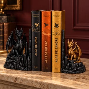 May include: Two dragon bookends featuring a black dragon on the left and a gold dragon on the right, both perched on jagged black rock bases. Between them are three books titled Alas de Onix, Alas de Hierro, and Alas de Sangre by Rebecca Yarros.
