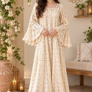 May include: A floor-length cream-colored dress featuring an intricate all-over crochet pattern, a scoop neckline, and long, wide bell sleeves with scalloped edges. The garment has a loose, flowing silhouette that extends to the floor.