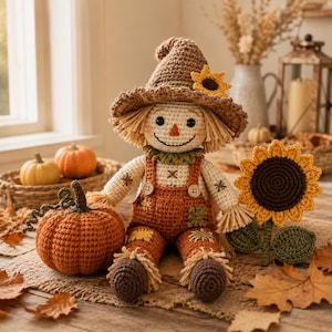 May include: A crocheted scarecrow doll wearing a brown hat with a yellow sunflower, a cream-colored long-sleeve shirt, and orange overalls with patches. The doll is positioned next to a crocheted orange pumpkin and a large crocheted sunflower.
