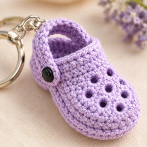 May include: A lavender crochet keyring shaped like a miniature clog shoe. The item features a textured, woven yarn construction with a series of small circular cutouts on the top and a single black circular button detail on the side strap.