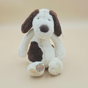 May include: A plush dog toy with cream-coloured fur, featuring dark brown floppy ears, a dark brown nose, and a large dark brown patch on its side. A small sticker with an apple graphic and the text "Happy Apple" is attached to the dog's foot.