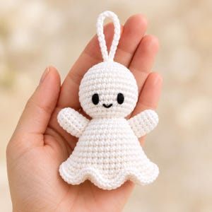 May include: A small white crocheted ornament shaped like a ghost or spirit. The figure has a round head, two small black embroidered eyes, a tiny black embroidered smile, and a flared skirt base. A white crocheted loop is attached to the top.