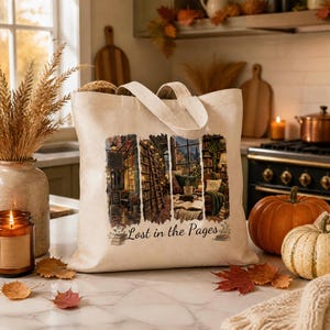May include: A beige canvas tote bag featuring a printed graphic of a cozy library interior with bookshelves, a ladder, and a window view. The text "Lost in the Pages" is printed below the image, accompanied by small illustrations of stacked books.