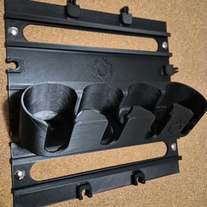 Peut inclure: Un organisateur mural noir imprimé en 3D doté d'une plaque arrière rectangulaire avec deux fentes horizontales et quatre supports circulaires intégrés. L'unité comprend de petits clips sur les bords supérieur et inférieur et un logo en forme d'engrenage.