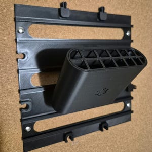 Peut inclure: Un organisateur mural noir imprimé en 3D composé d'une base rectangulaire avec des fentes horizontales et d'un support incliné en saillie doté d'une structure intérieure en grille. La base comporte de petites fixations métalliques.