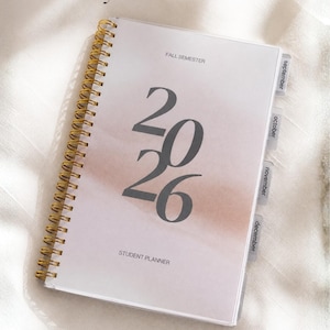 May include: A 5.5x8.5 inch spiral-bound student planner with a gold coil. The cover features a soft pink and white gradient background, the text "2026", "FALL SEMESTER", and "STUDENT PLANNER", with side tabs labelled September, October, November, and December.