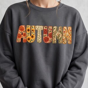 Peut inclure: Un sweat-shirt à col rond gris anthracite portant le mot AUTUMN imprimé sur la poitrine. Chaque lettre est remplie d'un motif botanique détaillé de fleurs orange et rouges, de tournesols jaunes, de fougères vertes et d'épis de blé sur fond crème.