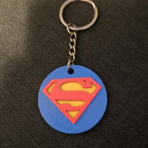 Puede incluir: Un llavero circular azul con un emblema en relieve rojo y amarillo del logotipo de Superman en el centro. El emblema está unido a una anilla de metal plateado mediante una cadena corta. La superficie del disco azul tiene una textura visible.