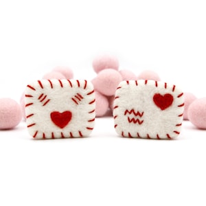 Op de afbeelding: Twee witte vilt envelopvormen met rode stiksels en rode vilt harten. Eén envelop heeft de letters "MM" erop gestikt.