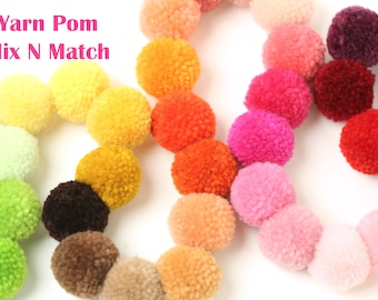 Può includere: Una collezione di pompon di lana colorati in varie tonalità di blu, verde, giallo, arancione, rosso, rosa, viola e marrone. I pompon sono disposti in cerchio, creando un effetto arcobaleno. Yarn Pom Mix N Match.