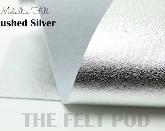 May include: Metallic felt fabric in brushed silver. The felt is folded over to show the back side, which is white. The text "Metallic Felt Brushed Silver" is printed at the top of the image. The text "THE FELT POD" is printed at the bottom of the image.