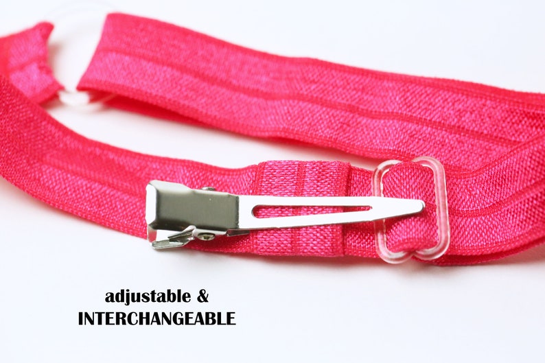 May include: A pink elastic headband with a silver clip and a clear plastic adjustable strap. The headband is adjustable and interchangeable. "adjustable & INTERCHANGEABLE" is printed on the image.