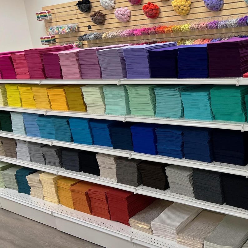 Puede incluir: Una pared de estantes llena de pilas de cuadrados de fieltro de colores en varios tonos de rojo, rosa, morado, azul, verde, amarillo, naranja, marr&oacute;n, gris y blanco.