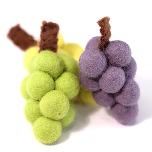 Puede incluir: Dos racimos de uvas de fieltro, uno verde y otro morado. Las uvas están hechas de bolas de fieltro y tienen tallos de fieltro marrón.