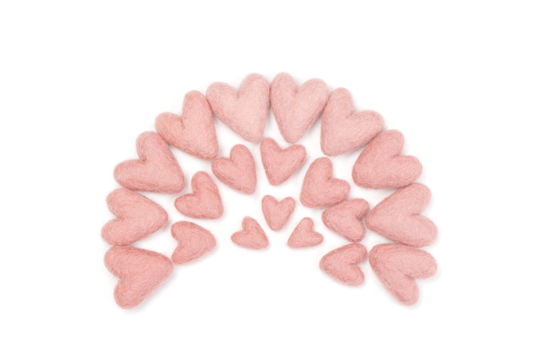 May include: A collection of pink felt hearts arranged in a semi-circle shape. The hearts are various sizes and are all a light shade of pink.