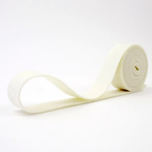 May include: A roll of off-white felt ribbon, unspooled and lying on a white background. The ribbon is soft and flexible, with a slightly textured surface. The roll is tightly wound, and the ribbon extends outward in a gentle curve.