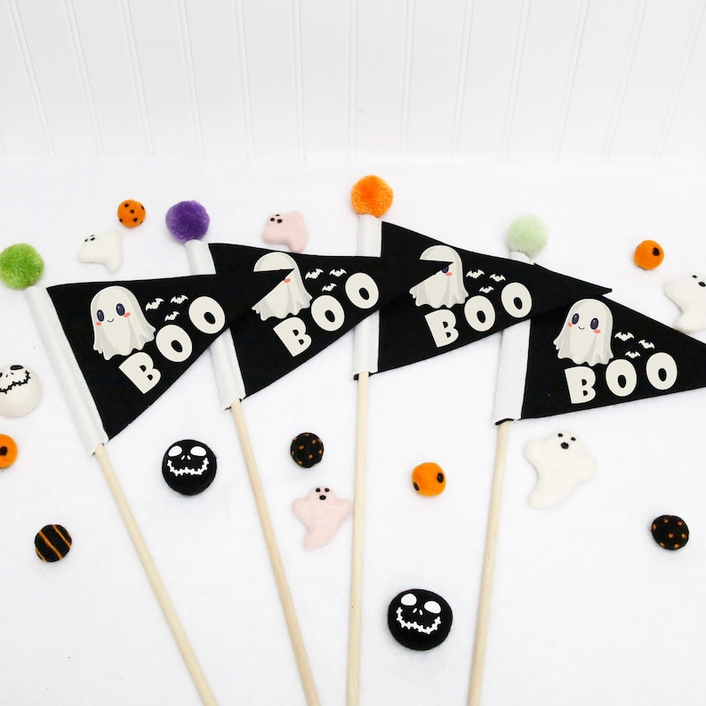 Op de afbeelding: Vier zwarte vlaggen met witte geestillustraties en het woord "BOO" erop gedrukt. De vlaggen zijn bevestigd aan houten stokken met kleurrijke pompoenen aan de bovenkant.