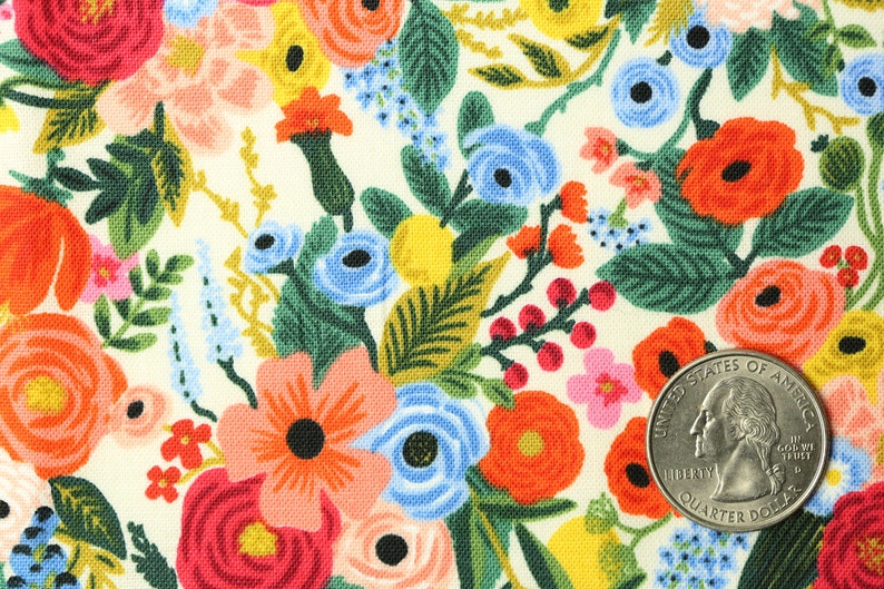 May include: Fabric with a floral pattern on a cream background. The design features a variety of flowers in red, orange, pink, blue, and yellow, with green foliage. A quarter coin is included for scale.