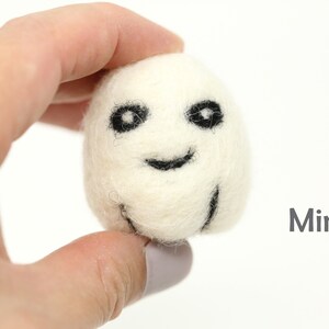 May include: A small white felt ghost with black eyes and a smiling mouth. The ghost is being held in a hand. The word "Mini" is written in gray text below the ghost.