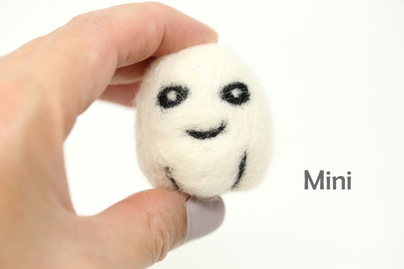 May include: A small white felt ghost with black eyes and a smiling mouth. The ghost is being held in a hand. The word "Mini" is written in gray text below the ghost.
