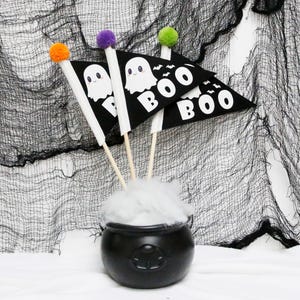 Op de afbeelding: Drie zwarte driehoekige vlaggen met witte geesten en "BOO"-ontwerpen, bevestigd aan houten stokken met oranje, paarse en groene pompoenen. De vlaggen zijn gerangschikt in een ketel met witte watten.