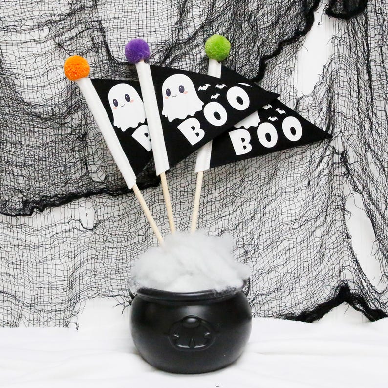 Op de afbeelding: Drie zwarte driehoekige vlaggen met witte geesten en "BOO"-ontwerpen, bevestigd aan houten stokken met oranje, paarse en groene pompoenen. De vlaggen zijn gerangschikt in een ketel met witte watten.