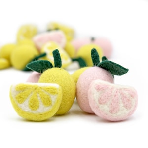 May include: A selection of felted lemons and pink citrus fruits with green leaves. The lemons are yellow with white detailing, and the citrus fruits are pink with white detailing. The fruits are arranged on a white background.