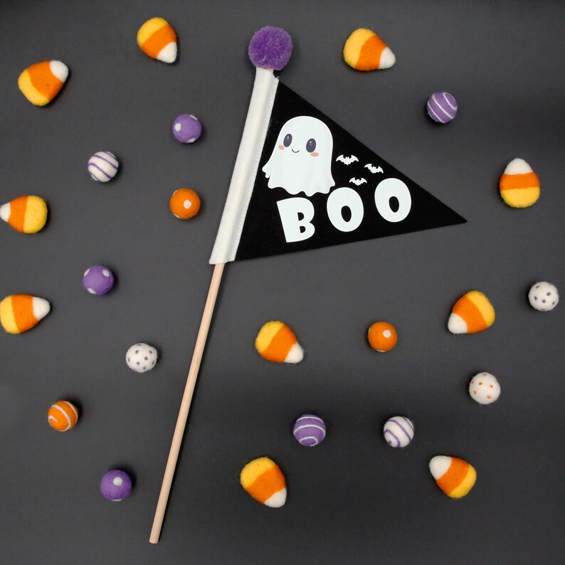 Op de afbeelding: Een zwarte vlag van vilt met een witte geest en het woord "BOO" erop. De vlag is bevestigd aan een houten stok en omringd door paarse en oranje ma&iuml;s snoepjes.