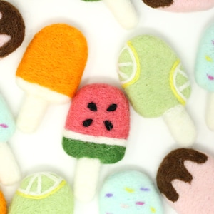 Puede incluir: Una colección de paletas de hielo de fieltro en varios sabores, incluyendo sandía, lima, naranja, chocolate y frambuesa azul. Cada paleta de hielo tiene un palo blanco y está decorada con chispas o chocolate.