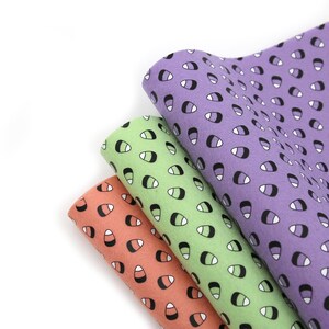 May include: Three rolls of fabric in coral, green, and purple, each patterned with a repeating candy corn design. The candy corn shapes are black and white on a colored background. The fabric is soft and appears to be felt.