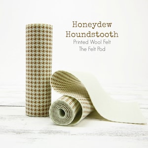 May include: Two rolls of brown and white houndstooth patterned wool felt. One roll is unrolled, showing the light green felt underneath. The text "Honeydew Houndstooth Printed Wool Felt The Felt Pod" is visible above the rolls.