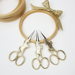 May include: Three gold unicorn-shaped embroidery scissors with silver blades. The scissors are arranged on a white surface with a wooden embroidery hoop in the background.