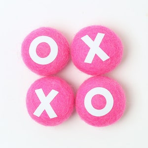May include: Four pink felt balls with white letters spelling out "XOXO" on a white background.