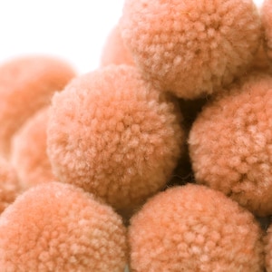 May include: A pile of soft, peach-colored pom poms. The pom poms are made of yarn and are fluffy and round.