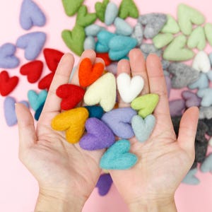 May include: A collection of felt hearts in various colors, including red, yellow, blue, green, purple, and white. The hearts are arranged in a pile in the palm of a hand.