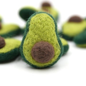 Puede incluir: Primer plano de varios adornos de aguacate de fieltro. Los aguacates son verdes y amarillos con una semilla marrón. El fieltro tiene un aspecto texturizado. El fondo es blanco y borroso.