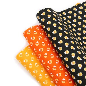 May include: Three rolls of Halloween-themed fabric in orange, yellow, and black. Each fabric features a repeating pattern of candy corn. The fabrics are made of a soft material and are suitable for various craft projects.