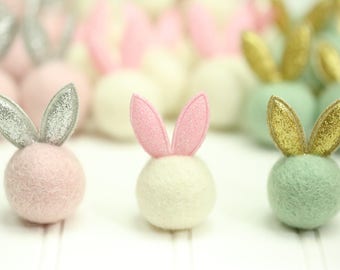 May include: Three felt balls with bunny ears. The balls are pink, white, and green. The bunny ears are silver, pink, and gold.